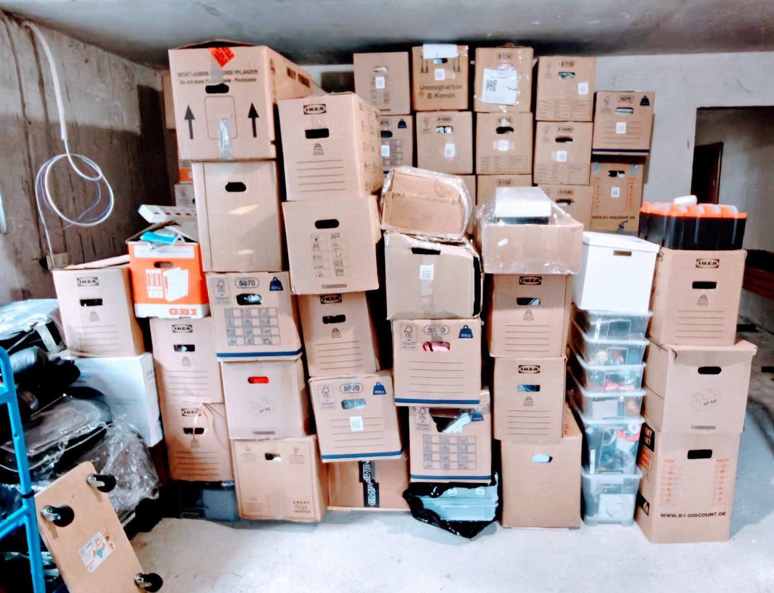 A storage room filled with stacked cardboard boxes of various sizes, some with clear plastic containers inside. The room has concrete walls and a concrete floor.