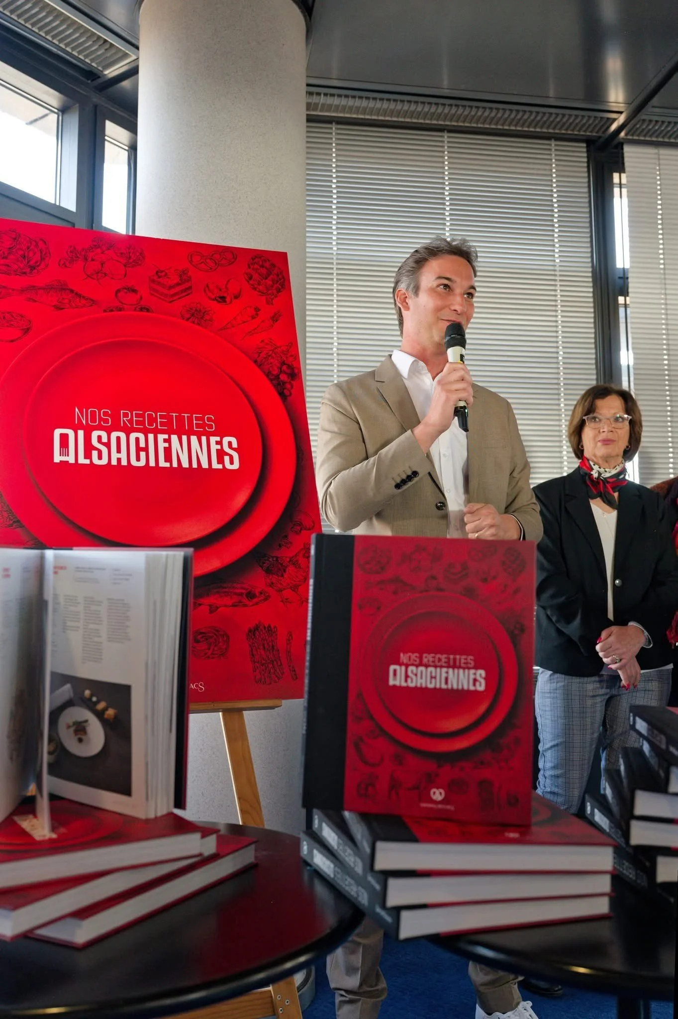 Une présentation de livre avec une affiche rouge et plusieurs exemplaires affichés en premier plan, un homme en costume clair parlant dans un micro, et une femme en costume noir avec un foulard rouge.