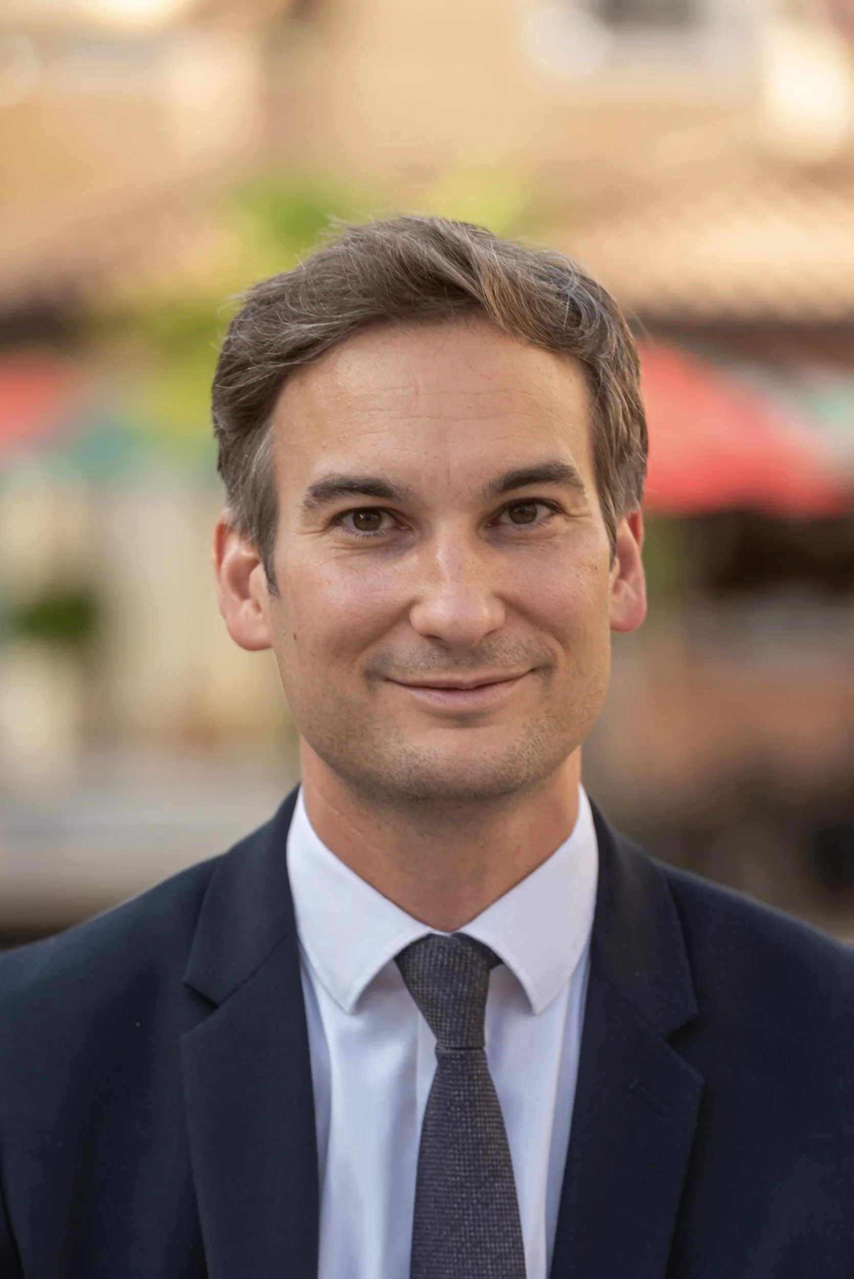 Un homme en costume et cravate posant dehors avec un arrière-plan flou.