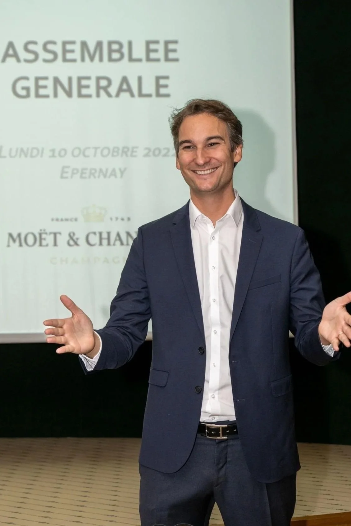 Un homme souriant en costume discutant lors d'une réunion d'entreprise, avec un écran derrière lui affichant 'Assemblée Générale' et des détails de date à Épernay.