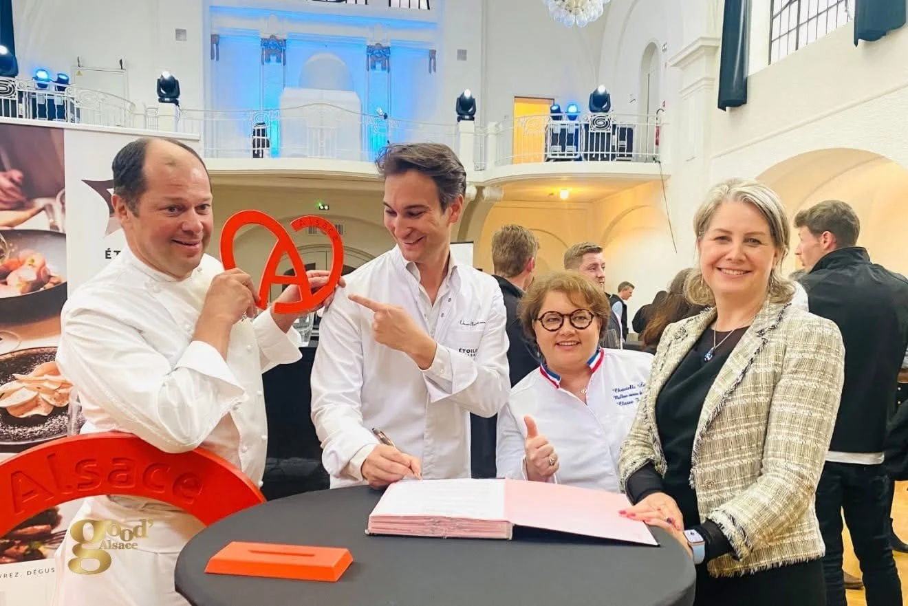 Groupe de quatre personnes souriantes lors d'un événement, tenant un symbole rouge avec le mot 'Alsace' visible, dans une grande salle bien éclairée avec d'autres personnes en arrière-plan.