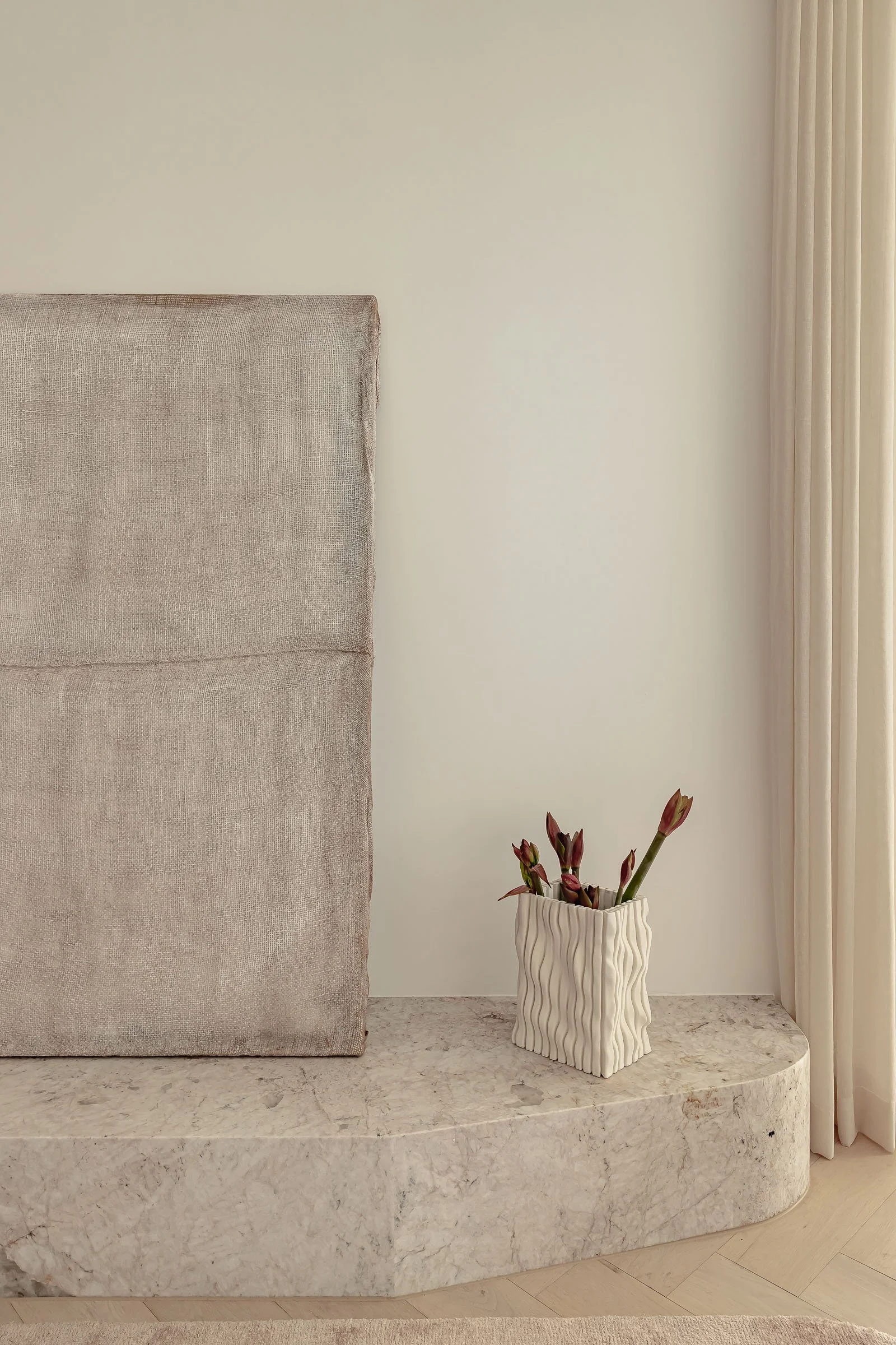 A beige upholstered headboard, a white textured planter with pinkish-red plants, a marble surface, and beige drapes in a minimalistic bedroom interior.