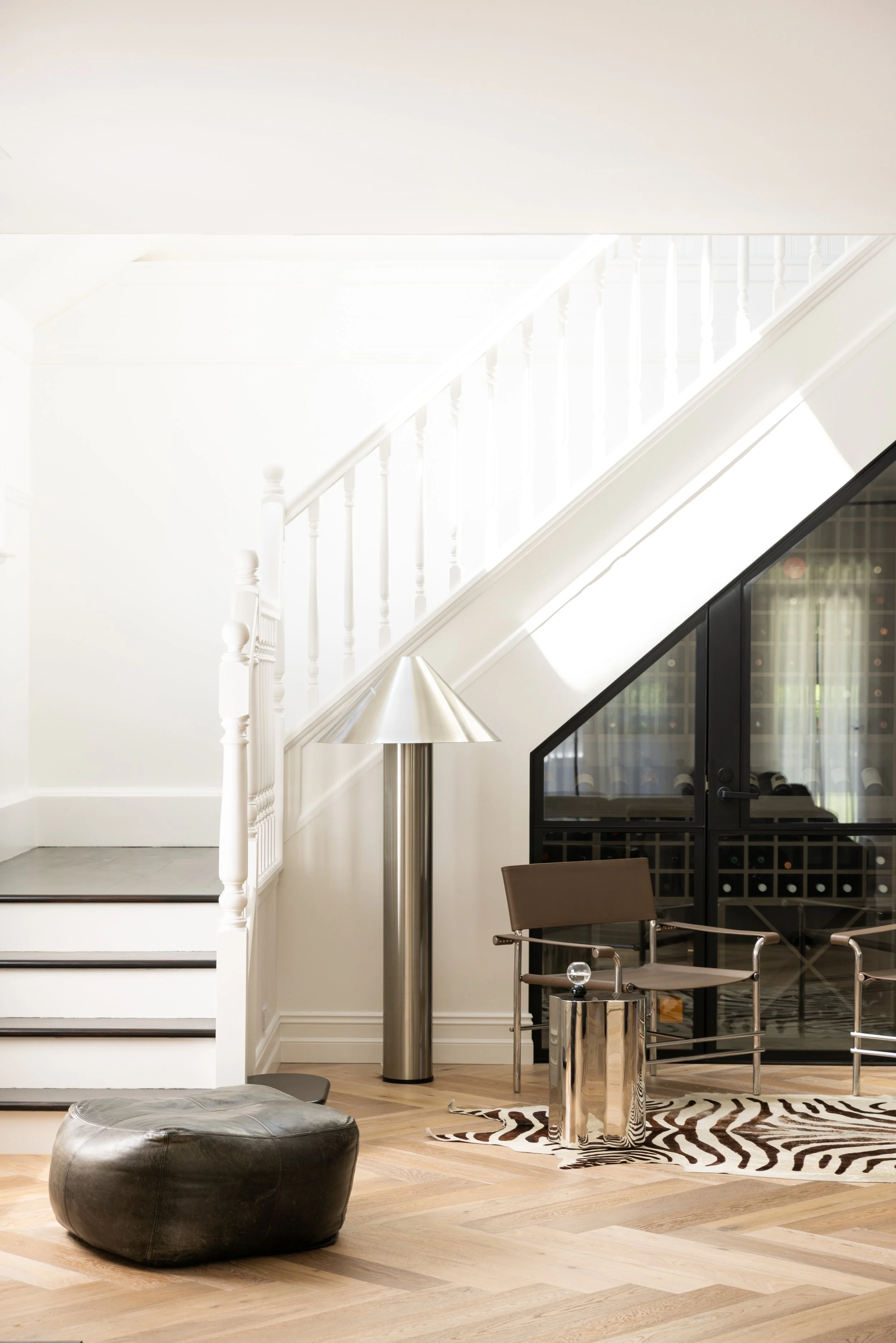Interior view of a modern home with a wooden staircase on the right side, a bedroom in the background with a bed covered in light-colored linens, and sunlight streaming through a large window with sheer curtains.