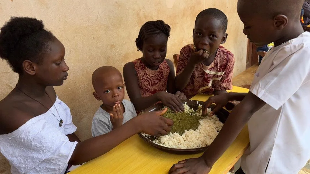 Children and a woman are gathered around a table with a large plate of food, which appears to be rice and vegetables, in a room with plain walls. One child is eating and others are touching the food.