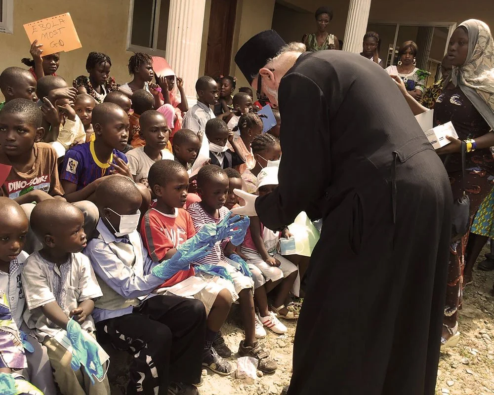 A man in a black robe and fez hat appears to be handling medical supplies or a vaccine to children and adults at an outdoor event with many children and women, some wearing masks and gloves.