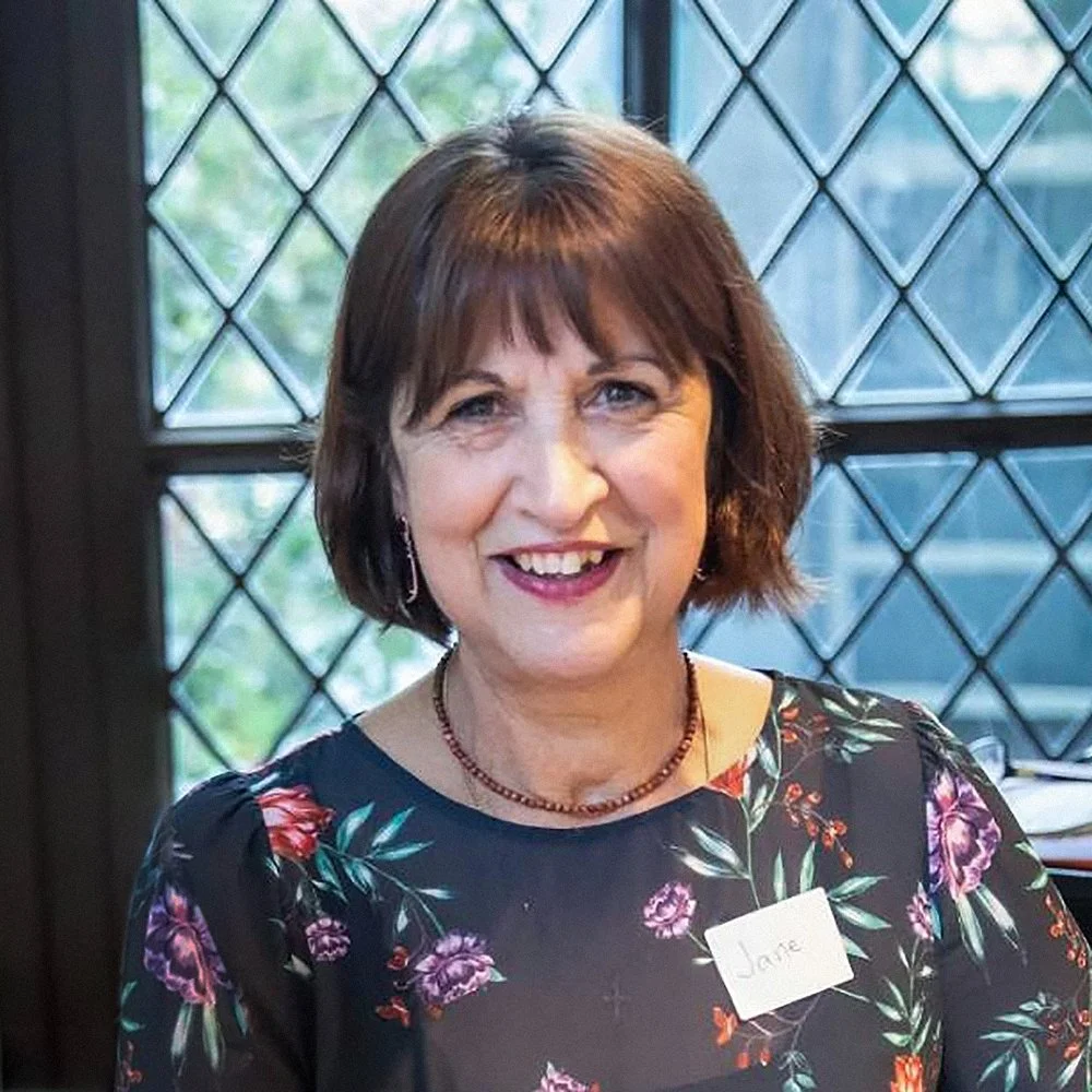 A woman with short brown hair, wearing a floral dress, is smiling at the camera. She has a name tag that says 'Jane' and is standing in front of a window with diamond-shaped glass panes.