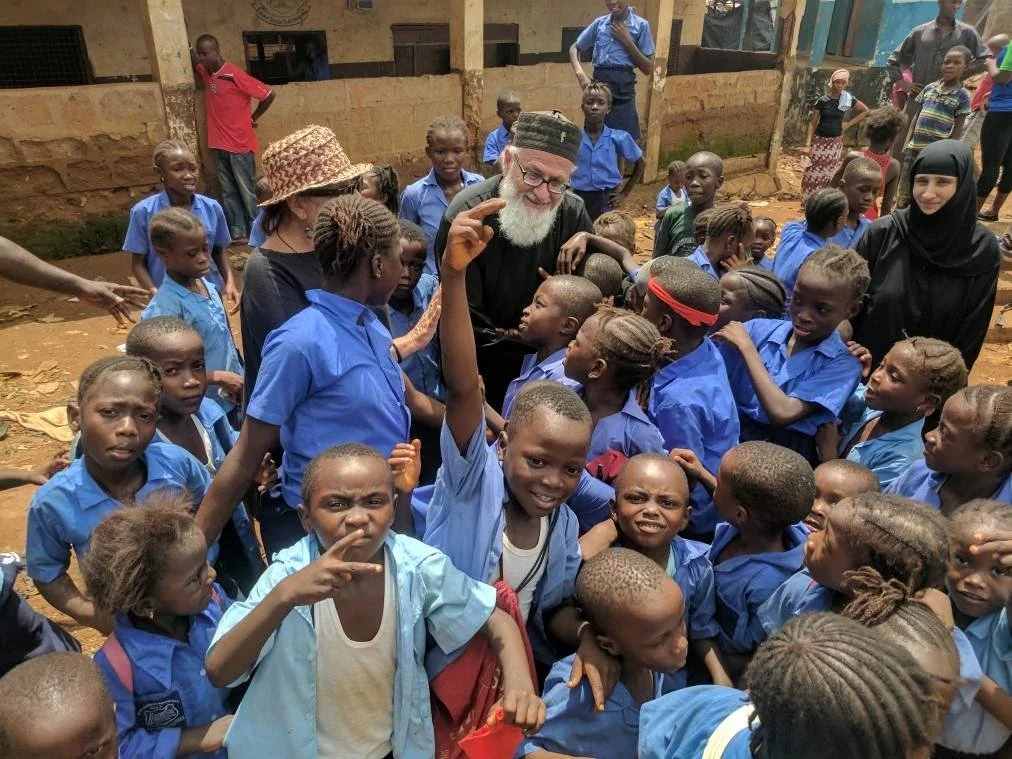 A group of children in blue school uniforms gathering around a smiling older man with glasses and a beard, who is talking to them. Several adults are present, including a woman in a black headscarf and a woman in a hat, in a rural outdoor setting.