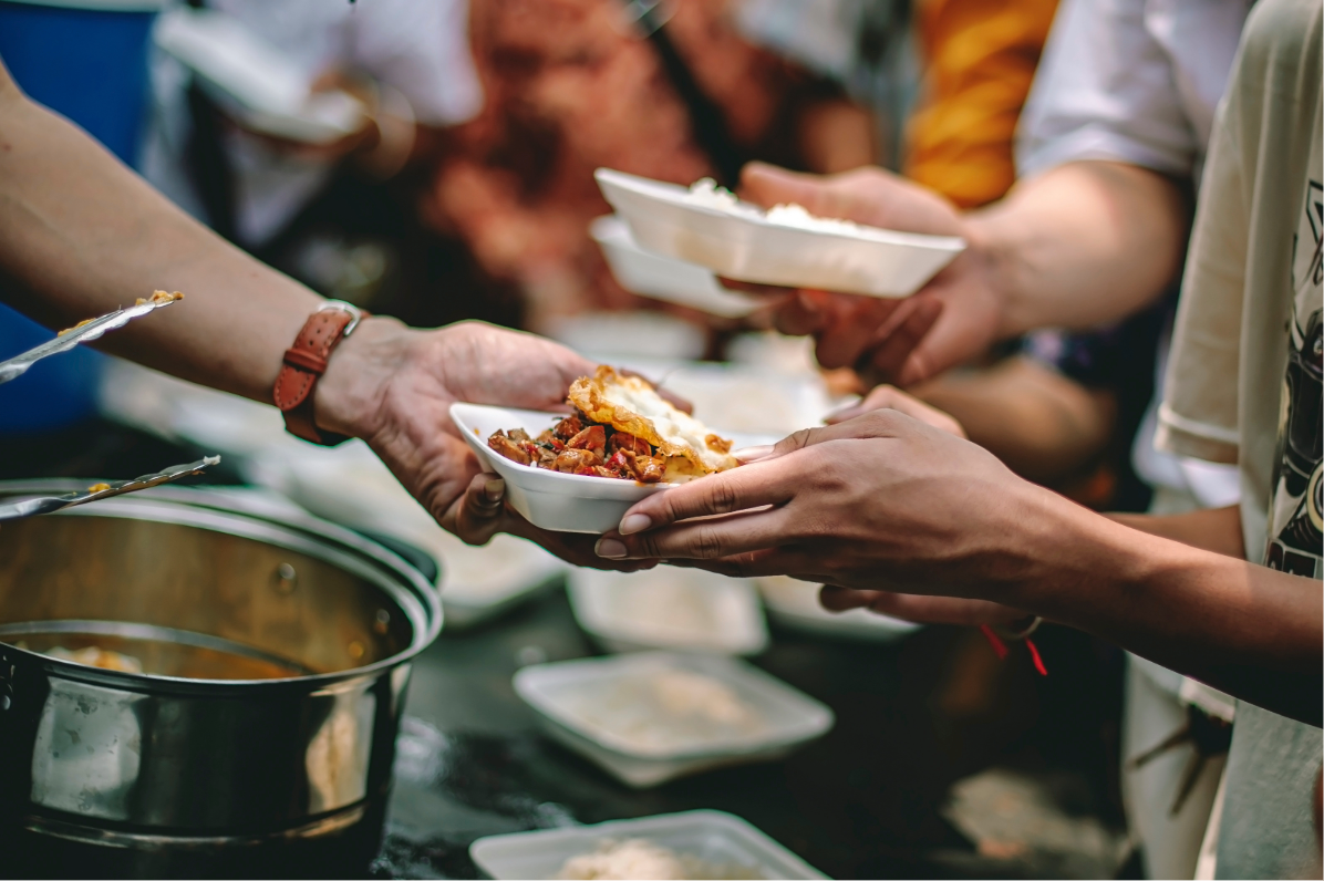 People serving and receiving food in a buffet line, with dishes and bowls on a table.
