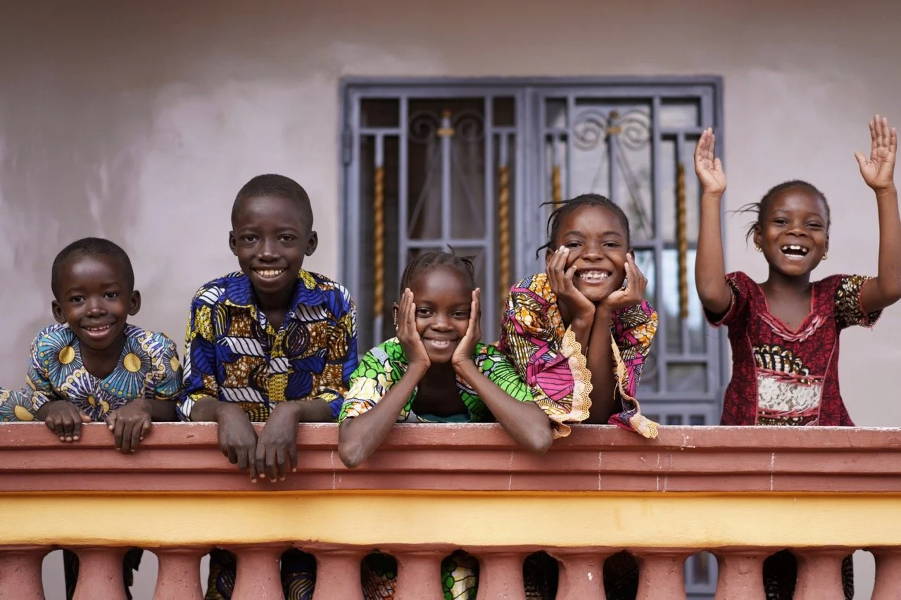 Five children of African descent standing behind a ledge, smiling and waving, wearing colorful traditional clothing, in front of a wall with a decorative metal window.
