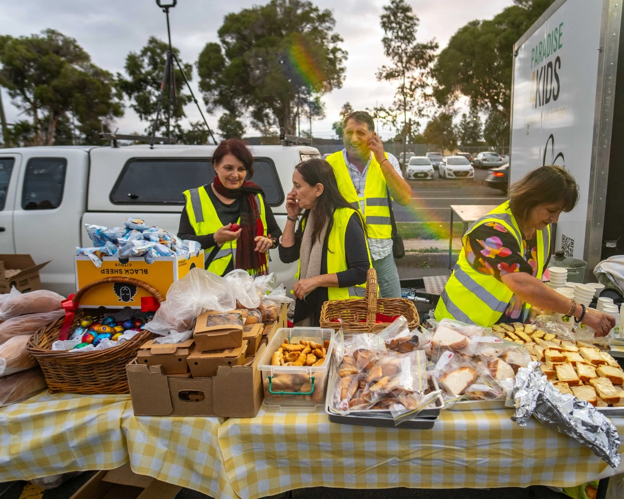 People wearing yellow safety vests standing around a table with snacks and baked goods, outdoors near a parking lot with trees in the background.
