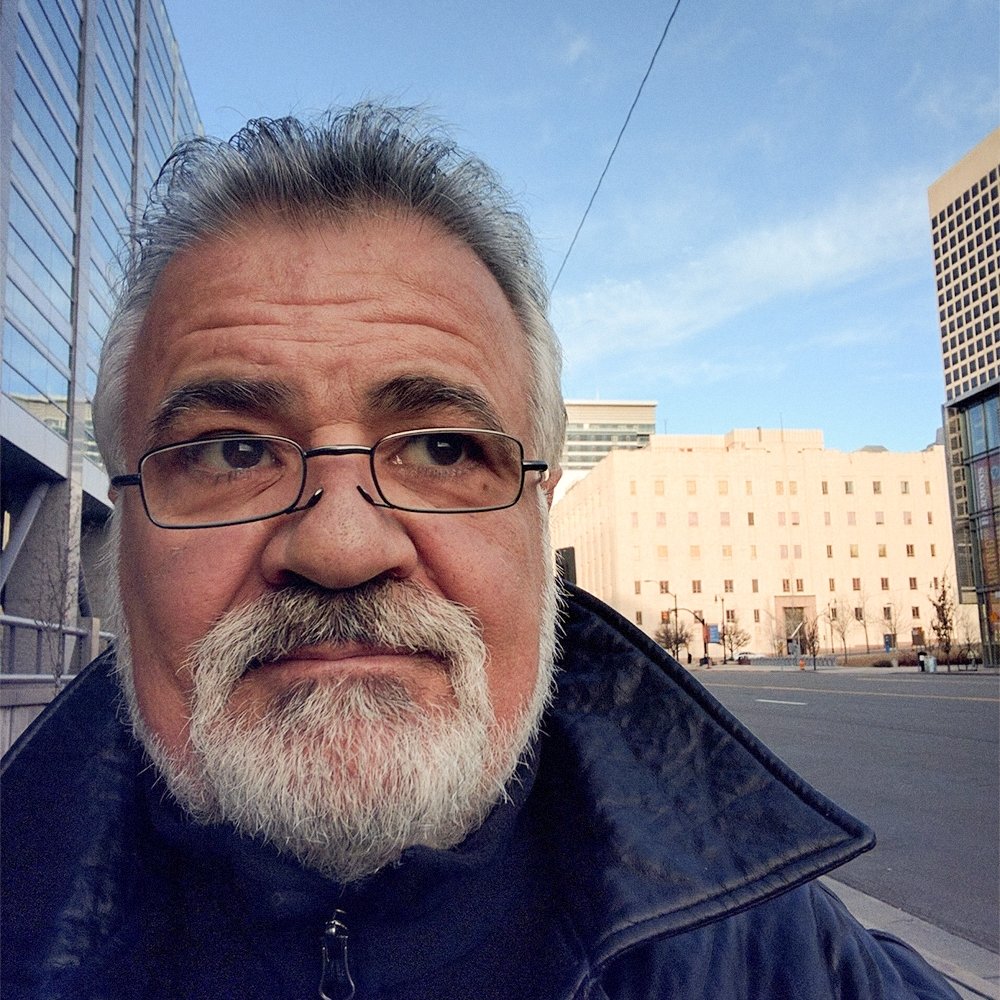 A close-up of a man with gray hair, a gray beard, and glasses, standing outside in an urban area with modern glass buildings and a city street in the background.