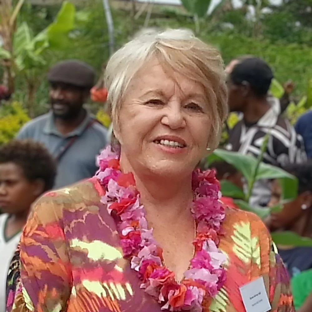 A smiling woman with short blonde hair wearing a colorful floral dress and a pink flower lei stands outdoors with greenery and other people in the background.