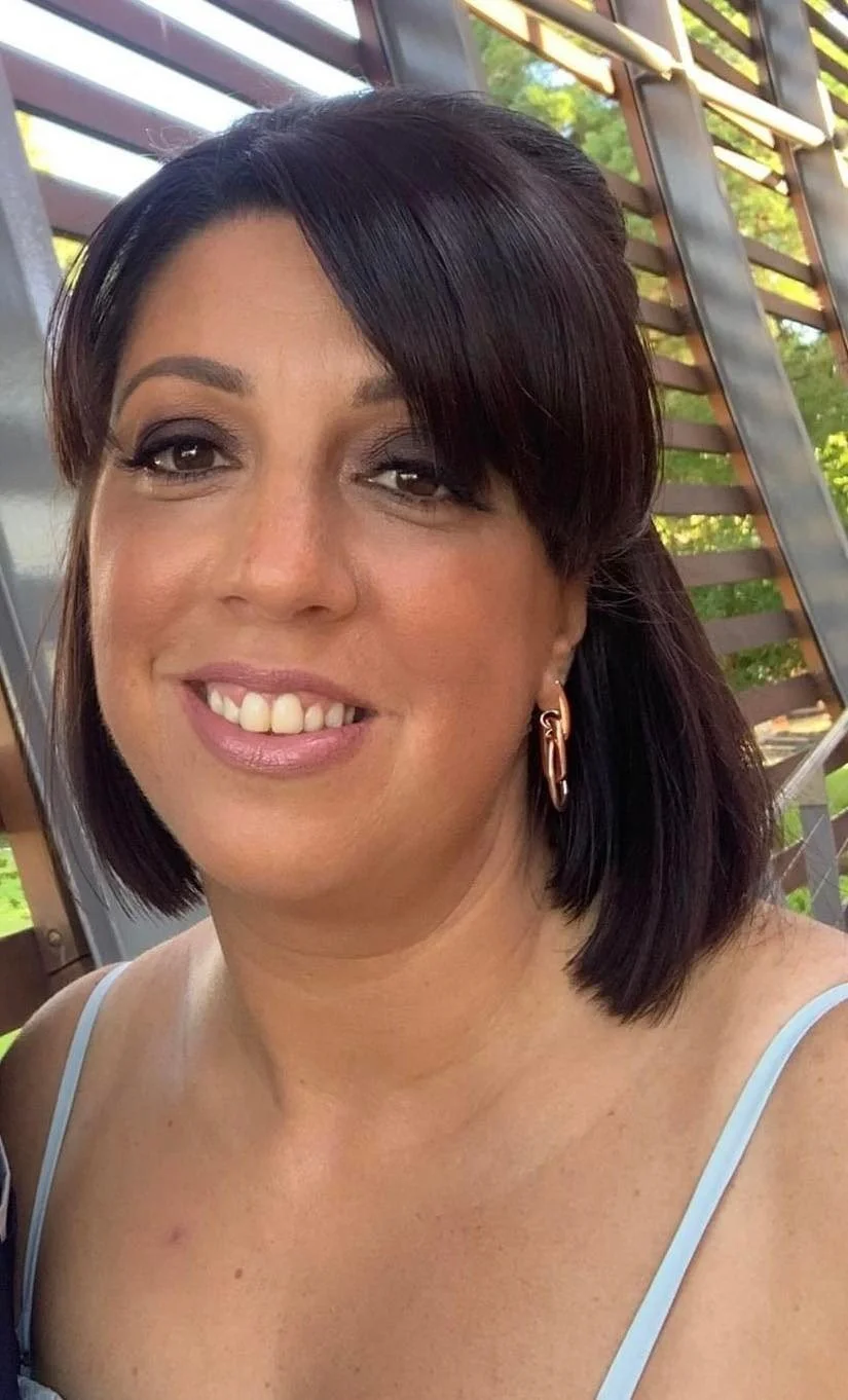 A woman with dark shoulder-length hair, wearing makeup, gold hoop earrings, and a light blue spaghetti strap top, smiling outdoors with a wooden and metal structure in the background.