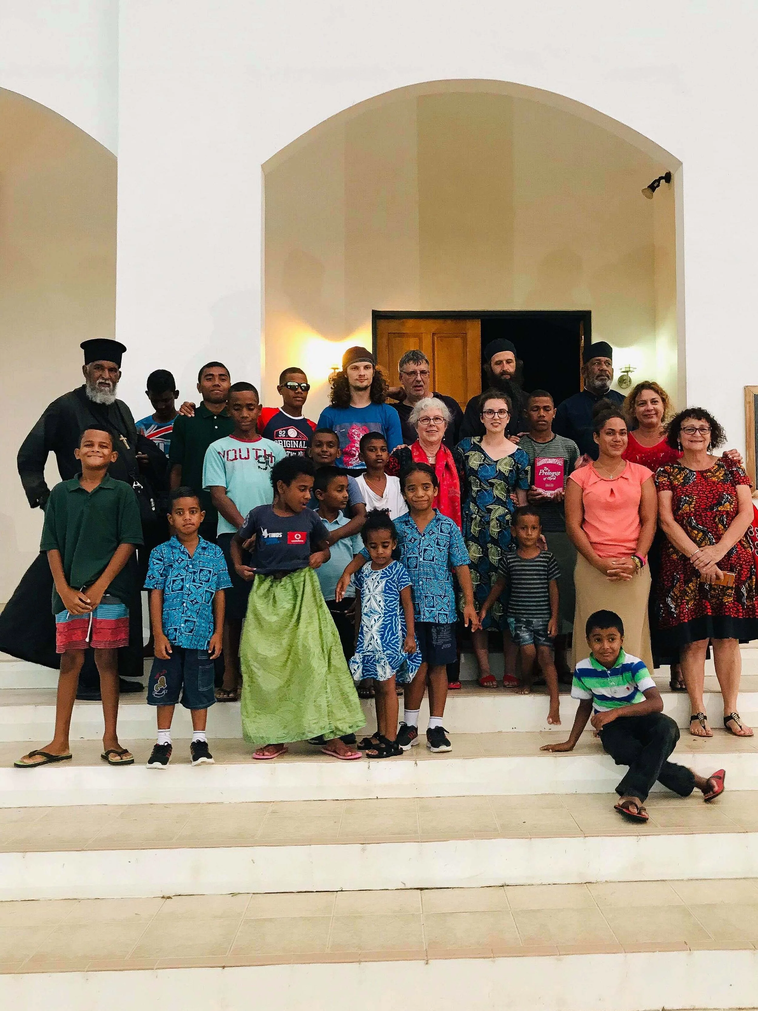 Group of children and adults standing on stairs inside a building, with some children dressed in traditional clothing and casual clothing, posing for a photo.