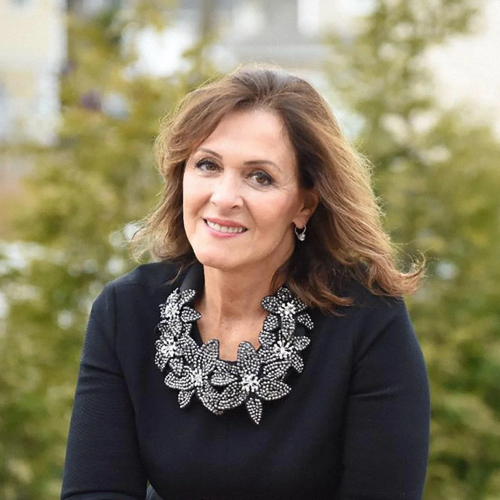 A woman with light skin and shoulder-length brown hair smiling outdoors. She is wearing a black top with a large, ornate, beaded necklace and matching earrings. The background is blurred with green foliage.