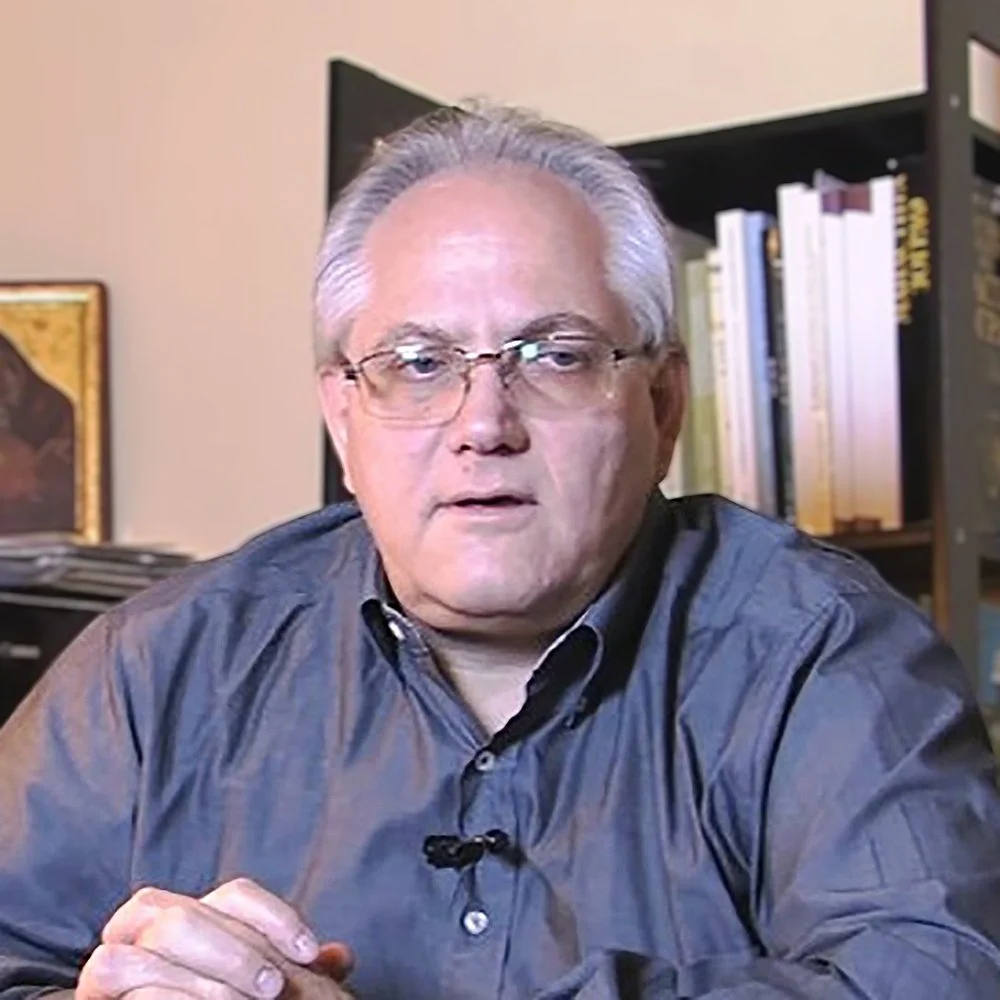 A light-skinned man with gray hair, glasses, and a serious expression, sitting in an office or study with bookshelves and framed artwork in the background.