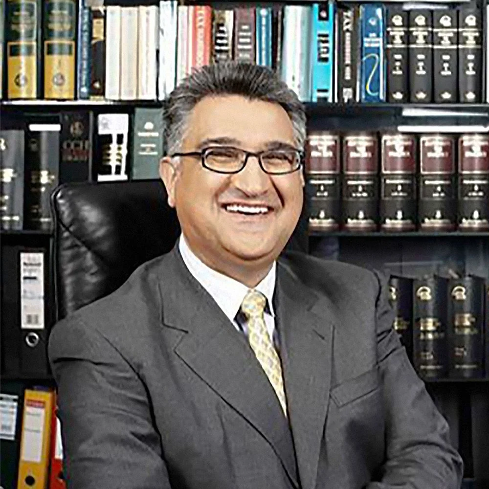 A man in a suit and tie sitting in an office with shelves of books and legal binders behind him, smiling at the camera.