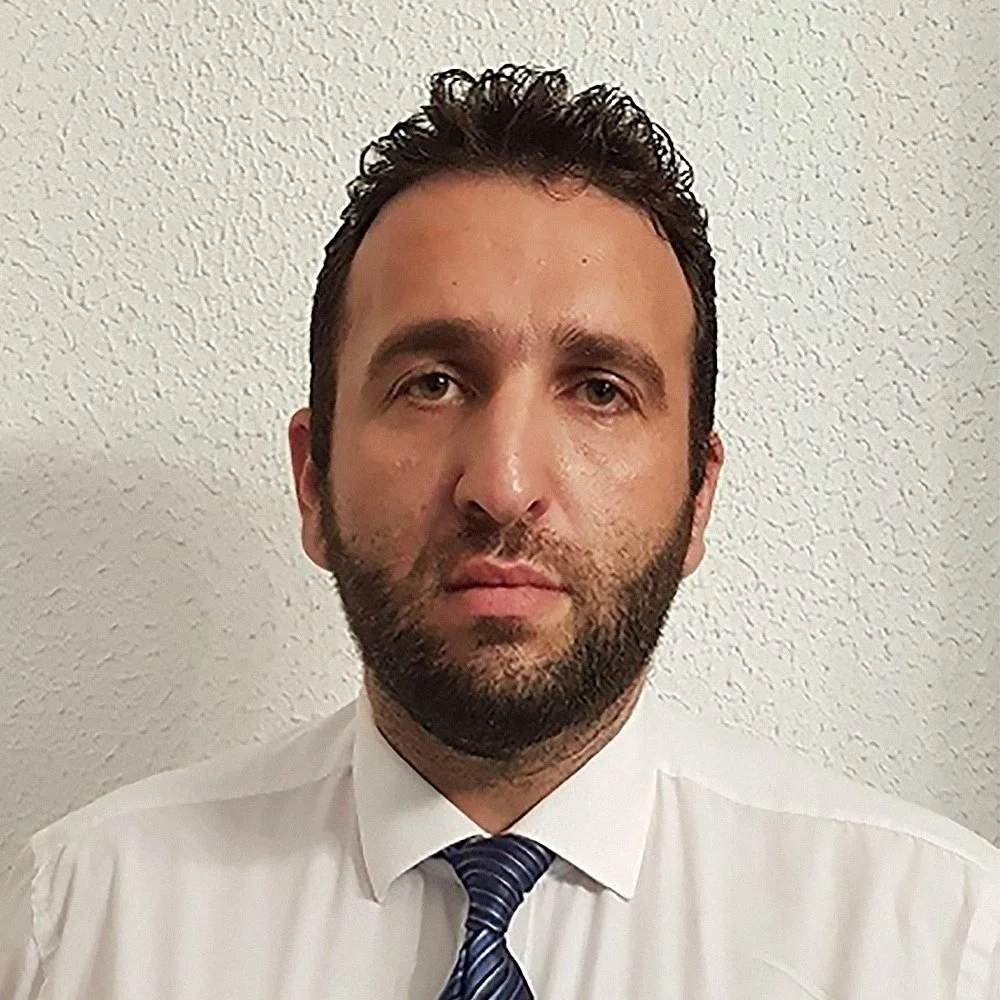 A man with dark, curly hair, a beard, wearing a white dress shirt and tie, standing against a textured off-white wall.