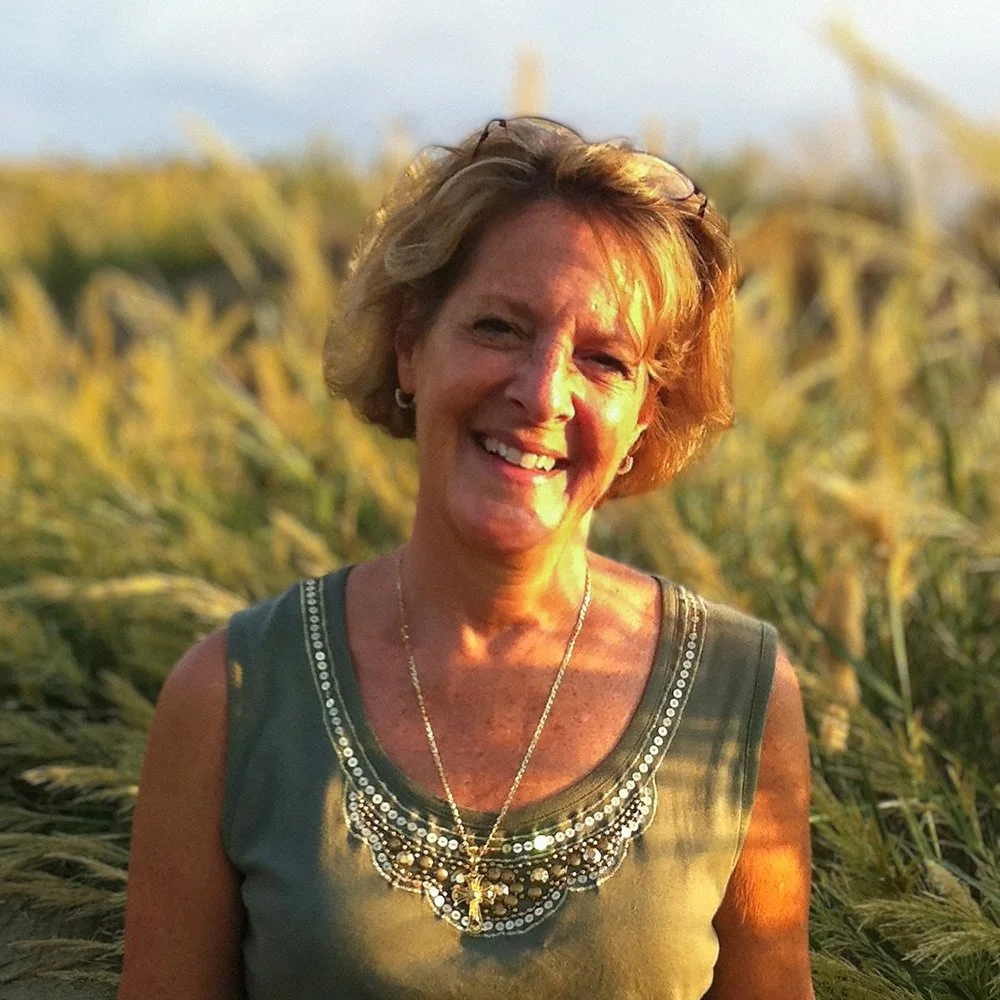 A smiling woman with short blonde hair wearing a sleeveless top and jewelry, standing in a field of tall grass at sunset.