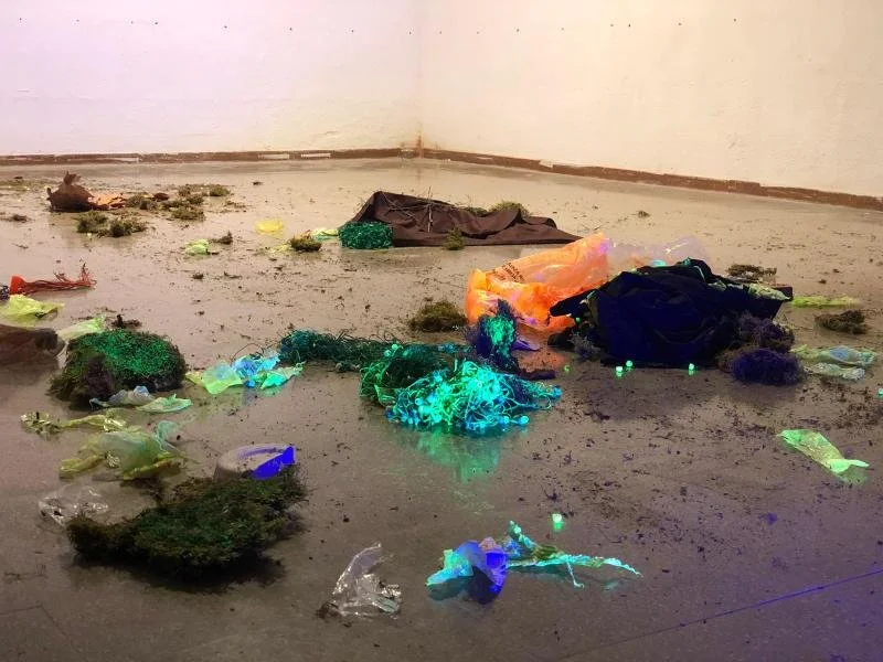 Arte conceptual con basura, luces y materiales diversos dispersos en un espacio interior con paredes blancas.