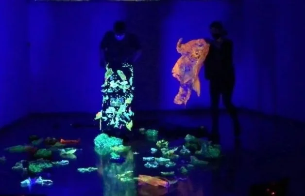 Dos personas observando obras de arte con iluminación negra y efectos fluorescentes en un espacio oscuro.