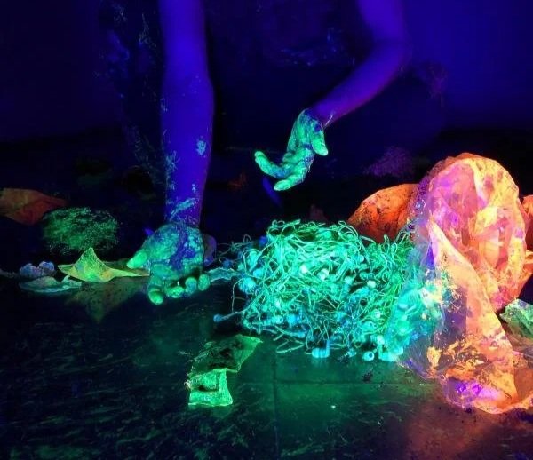 Mano sosteniendo una pila de cables de luces fluorescentes en un entorno oscuro con iluminación ultravioleta, y varias rocas y objetos con efectos de luz