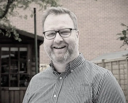 A man with glasses and a beard smiling outdoors, wearing a checkered button-up shirt, with a brick building and a wooden fence in the background.