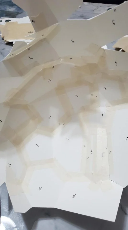An interior view of a precision-engineered paper sculpture by LUMISCA, showing multiple low-poly white paper planes numbered and secured with masking tape at calculated angles, forming a self-supporting structural network.