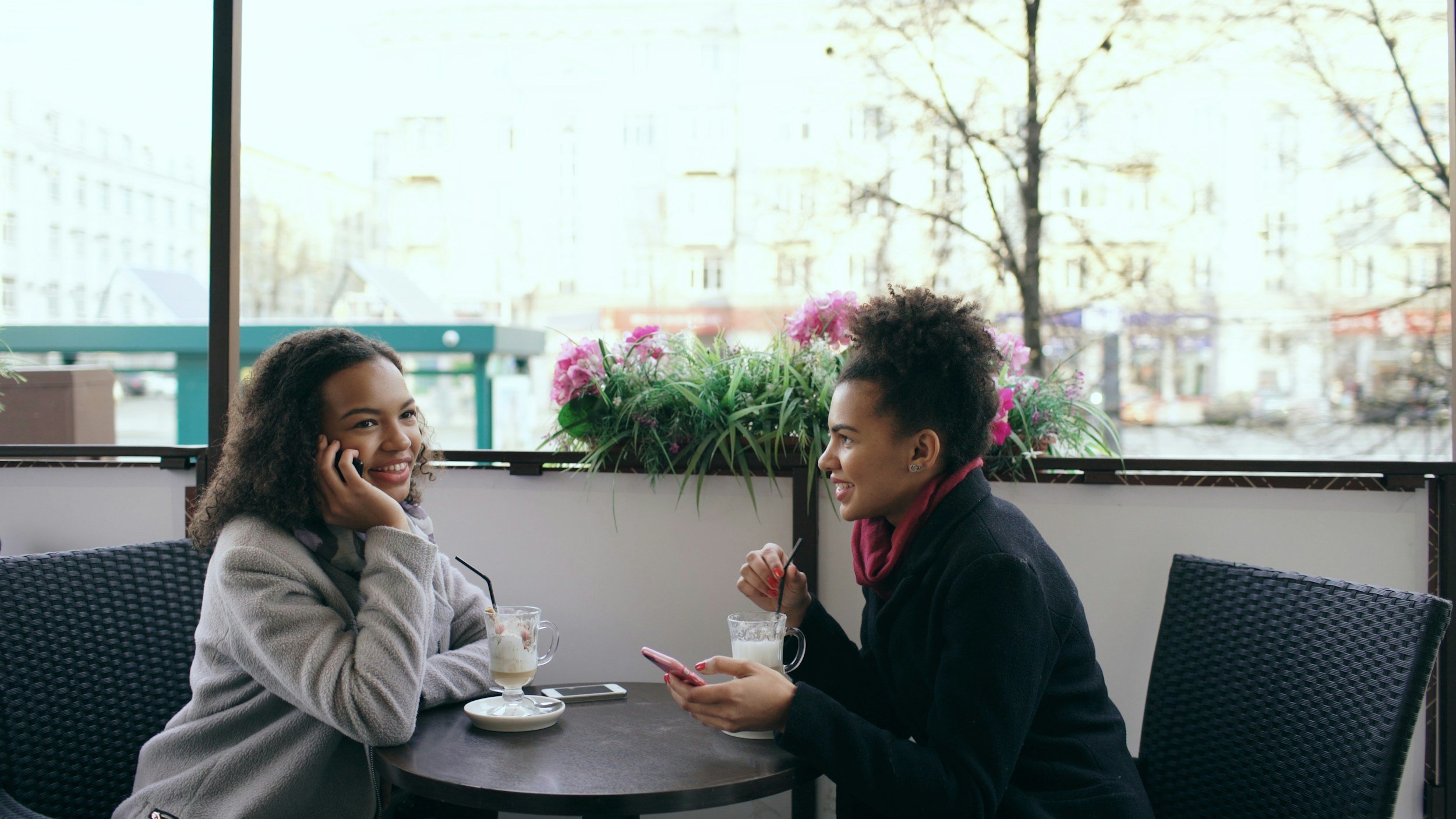 Two women sitting at a table in a bright cafe, smiling and talking with each other, with milkshakes in front of them and a large window showing trees in the background.