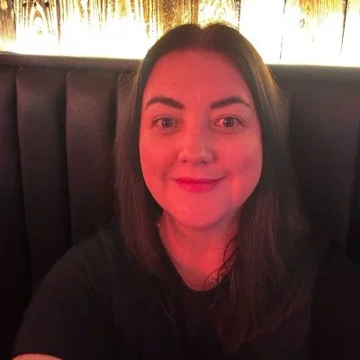 A woman with shoulder-length dark hair smiling at the camera in a dimly lit setting with a dark upholstered seat and a warm background.