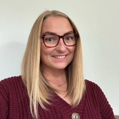 A smiling woman with blonde hair, wearing glasses and a maroon sweater, posing against a plain off-white background.