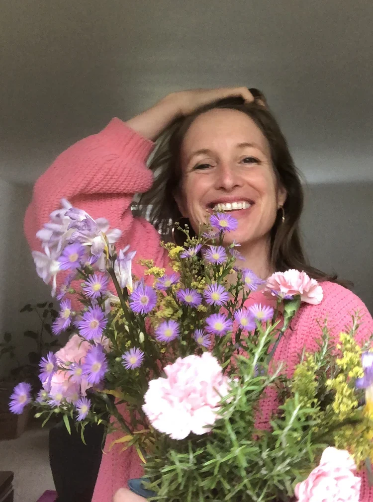 A woman smiling and holding a colorful bouquet of flowers while wearing a pink sweater.