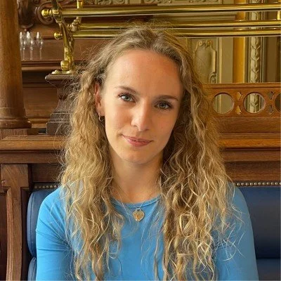 A woman with long, curly blonde hair wearing a blue top and a heart-shaped necklace, sitting indoors in front of a wooden and brass background.