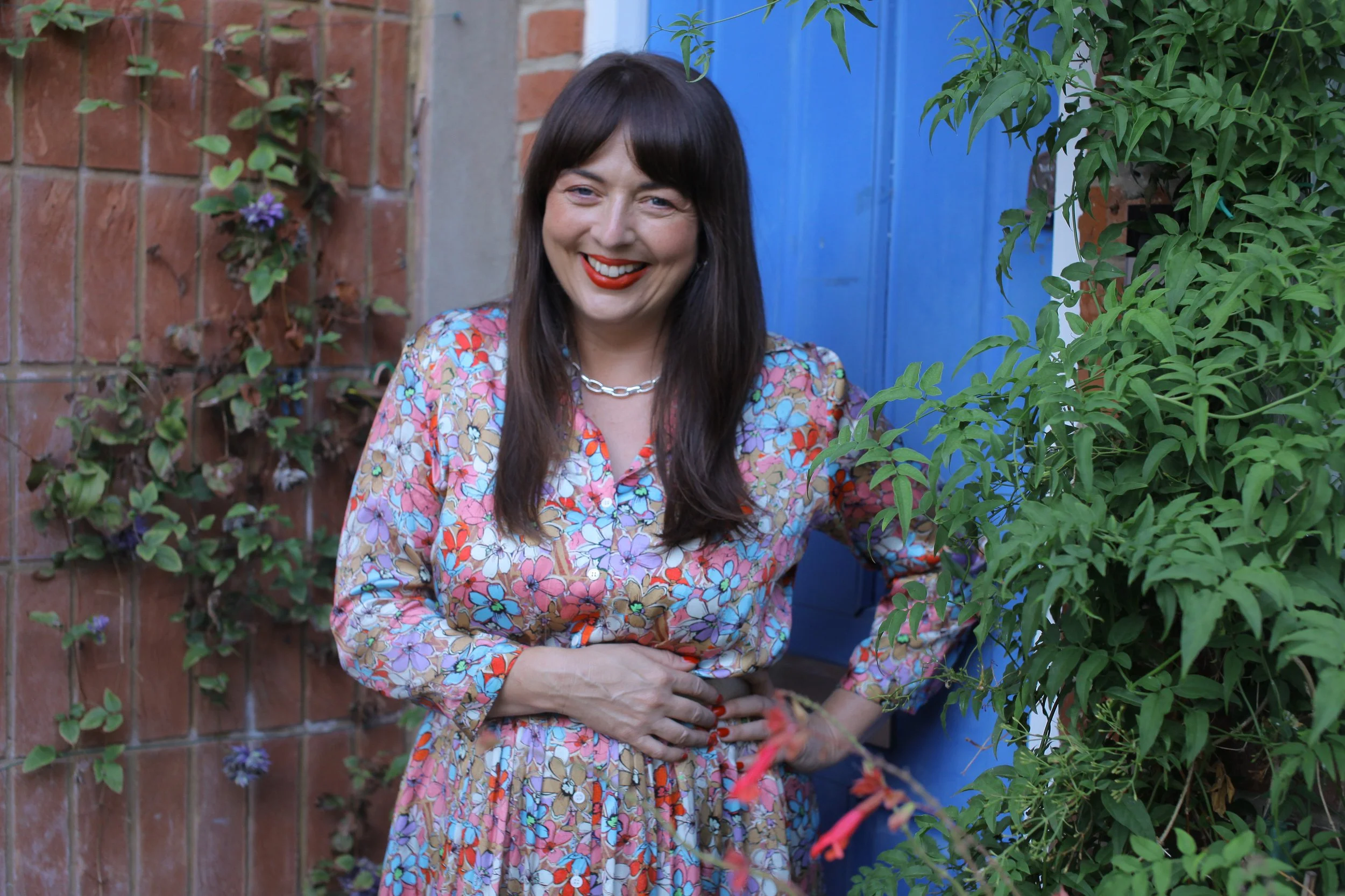 A woman with dark brown hair and red lipstick is standing outdoors, smiling. She is wearing a colorful floral dress and a silver chain necklace. The background features a brick wall with green ivy and a blue door or wall.