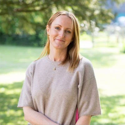 A woman with blonde hair smiling outdoors in a park or garden with green trees and grass in the background.