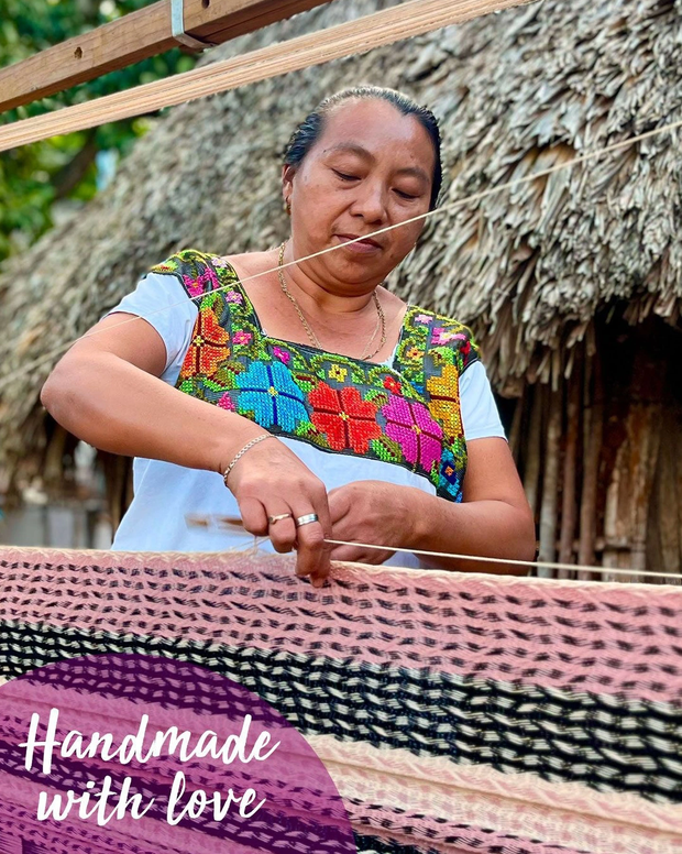 A woman in a traditional colorful embroidered blouse is hanging woven textiles on a line outdoors. She is focused on her work, with a thatched roof in the background. Text overlay says 'Handmade with love'.