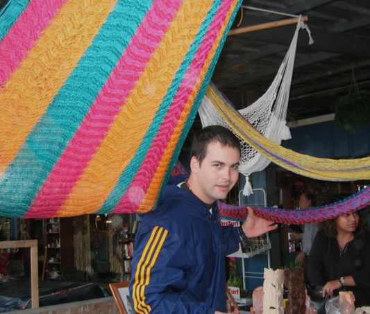 A young man in a blue jacket with yellow stripes is standing under colorful hanging decor at a market or fair.