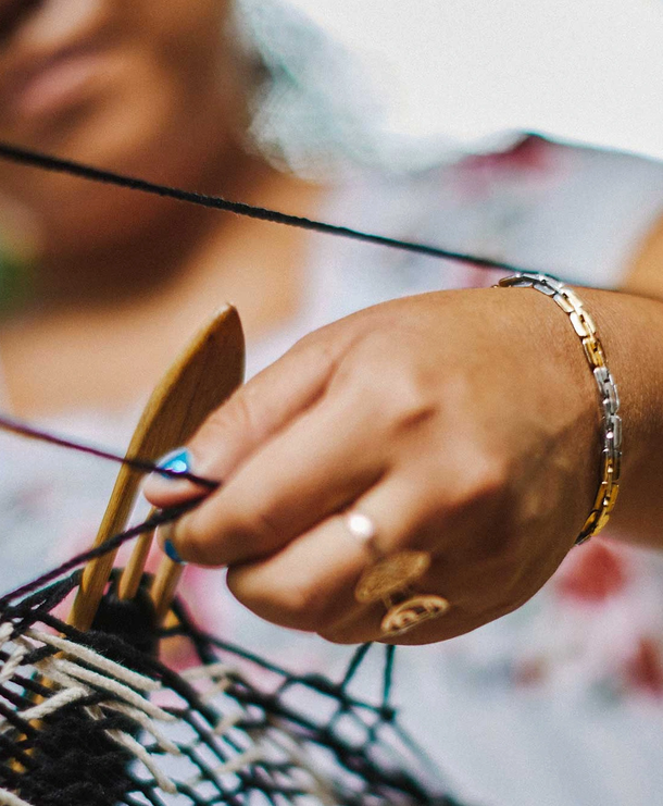 Person weaving with sticks, using their right hand, holding sticks with black and purple threads, wearing a gold and silver bracelet and multiple rings.