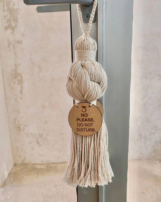 A beige tassel made of thick rope hangs from a hook on a metal frame, with a circular cardboard tag that reads 'NO PLEASE, DO NOT DISTURB.'