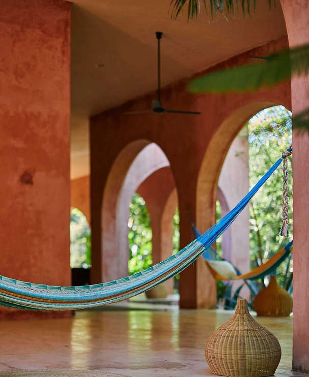 A porch with pinkish arches, a ceiling fan, and two blue hammocks hanging. A woven vase is placed on the floor, and there are trees visible through the arches.