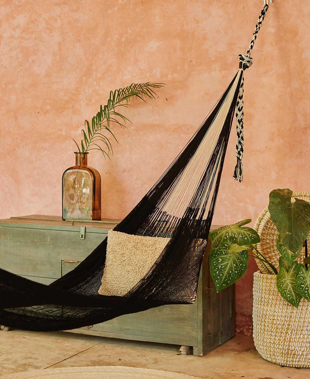 Indoor scene featuring a black and beige hammock hanging next to a light blue chest, a green potted plant with large leaves, and a taller plant in a brown vase on a pink textured wall.