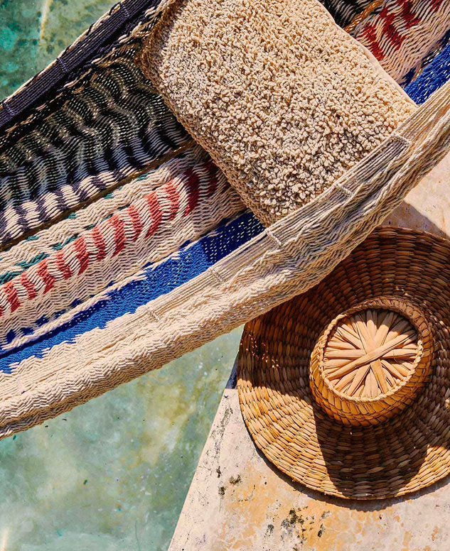 Beach scene with a colorful woven towel, a straw hat with a woven ornament, and clear water.