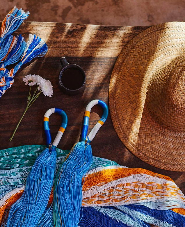 A straw hat, two blue and white towels, a black coffee mug, a daisy flower, and a bundle of blue, white, and orange yarns are arranged on a wooden surface with sunlight and shadows.