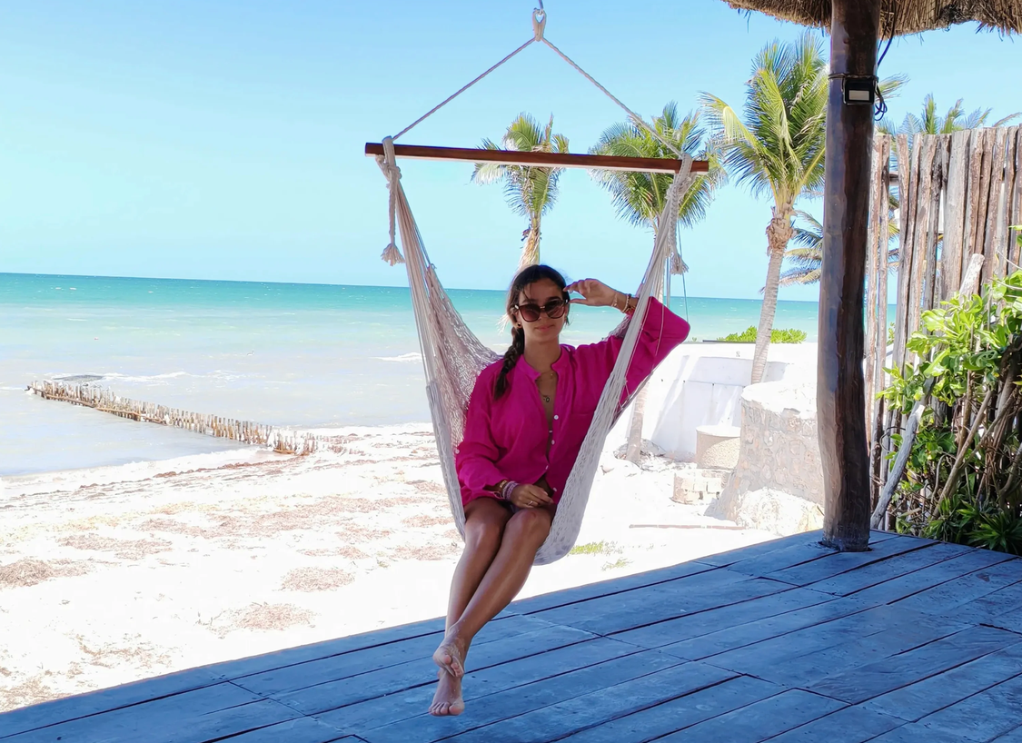 Woman in pink shirt sitting on a hanging hammock chair on a beach with palm trees, sandy shore, and ocean in the background.
