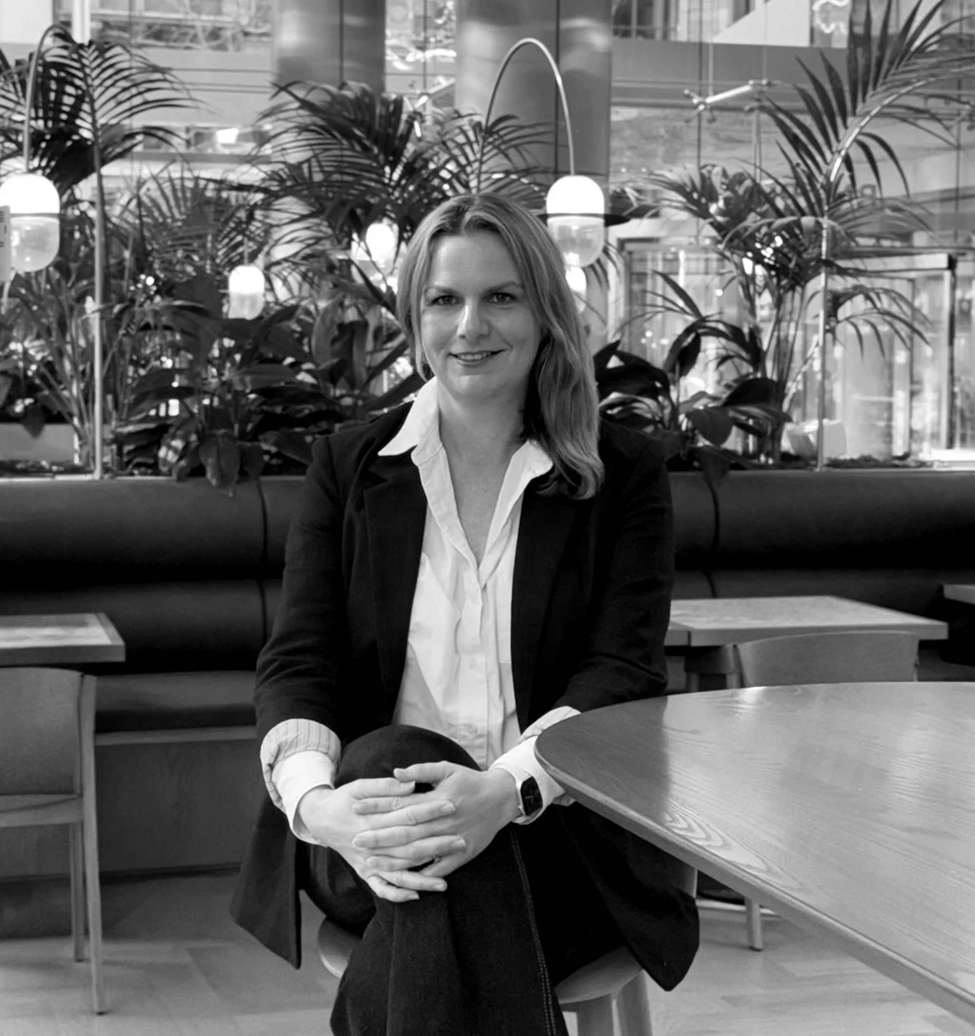 Black and white photo of Tessa Pawson with long hair, sitting at a table in a modern indoor space, surrounded by large leafy plants and spherical lamps, with glass walls and urban view in the background.