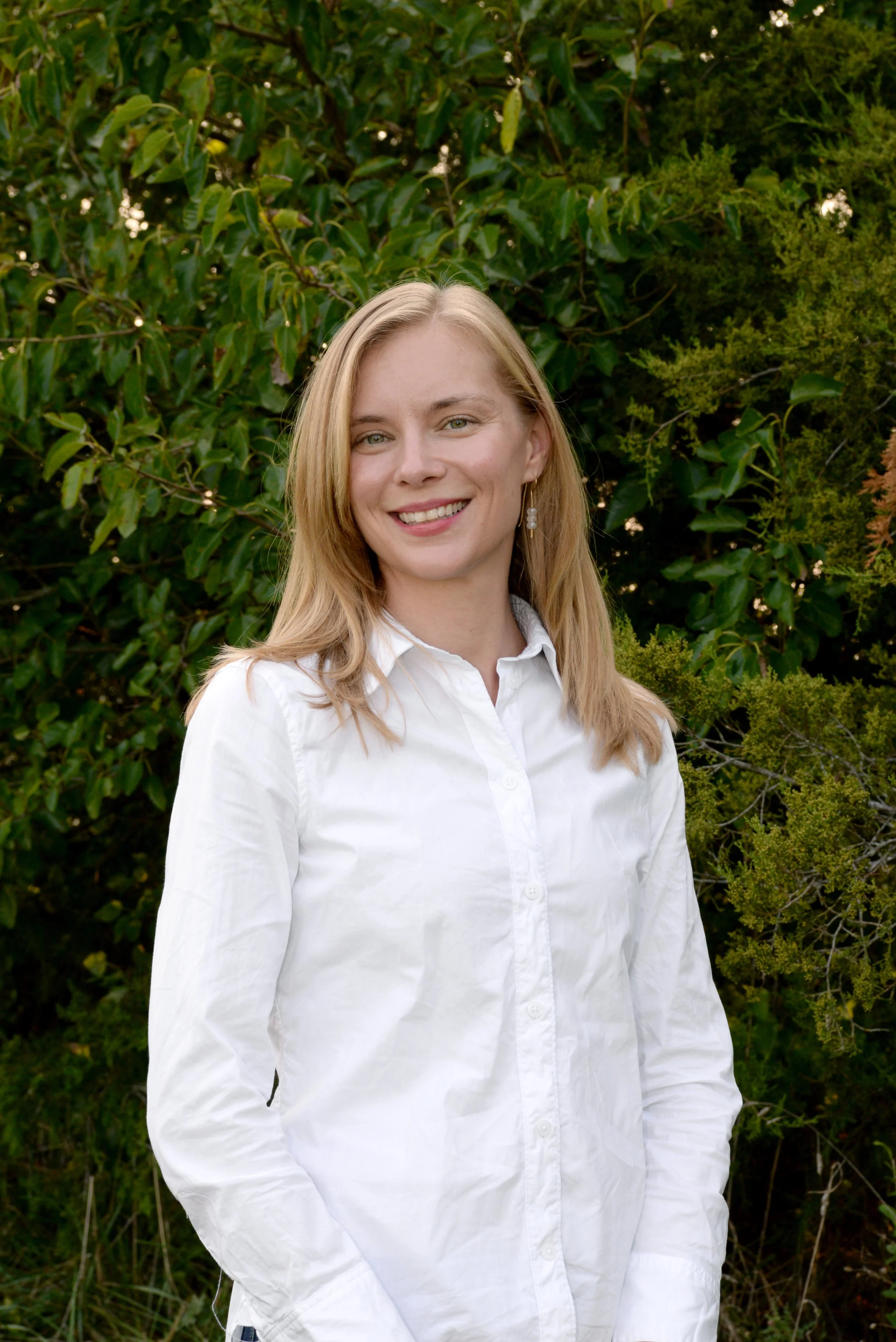 A woman with blonde hair, wearing a white button-up shirt, smiling outdoors in front of green foliage.