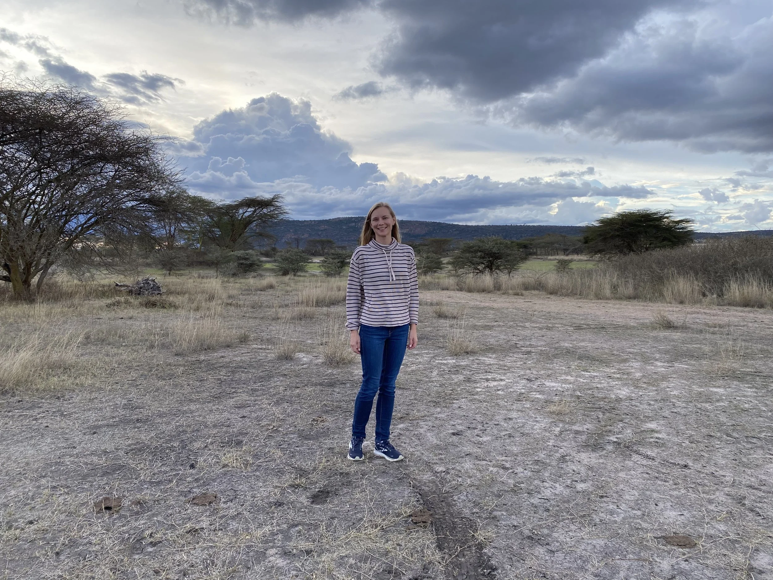 A woman standing on a dirt path in a savannah landscape with sparse trees and a cloudy sky.