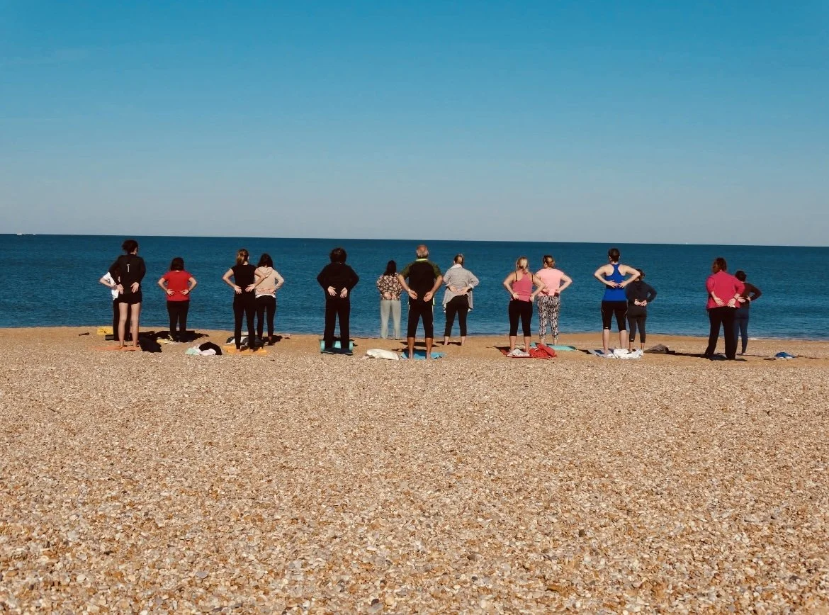 Groupe de personnes en tenue de sport, alignées sur la plage, participant à une séance de yoga ou de méditation face à la mer sous un ciel clair.