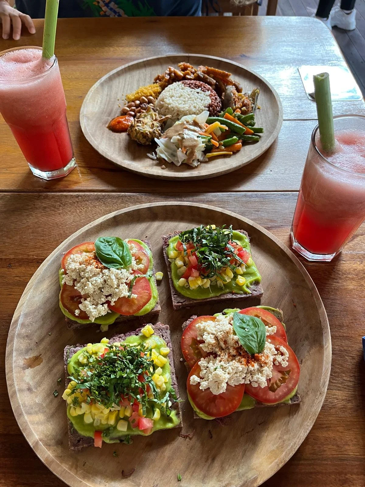 Deux assiettes avec des plats et des boissons. La première assiette contient des sandwichs avec des tomates, du fromage, de la salsa, et des herbes. La deuxième assiette contient un plat avec du riz, des légumes sautés, des haricots, et une portion de poulet. Deux verres de boisson rose avec des pailles en bois sont placés à côté des assiettes sur une table en bois.