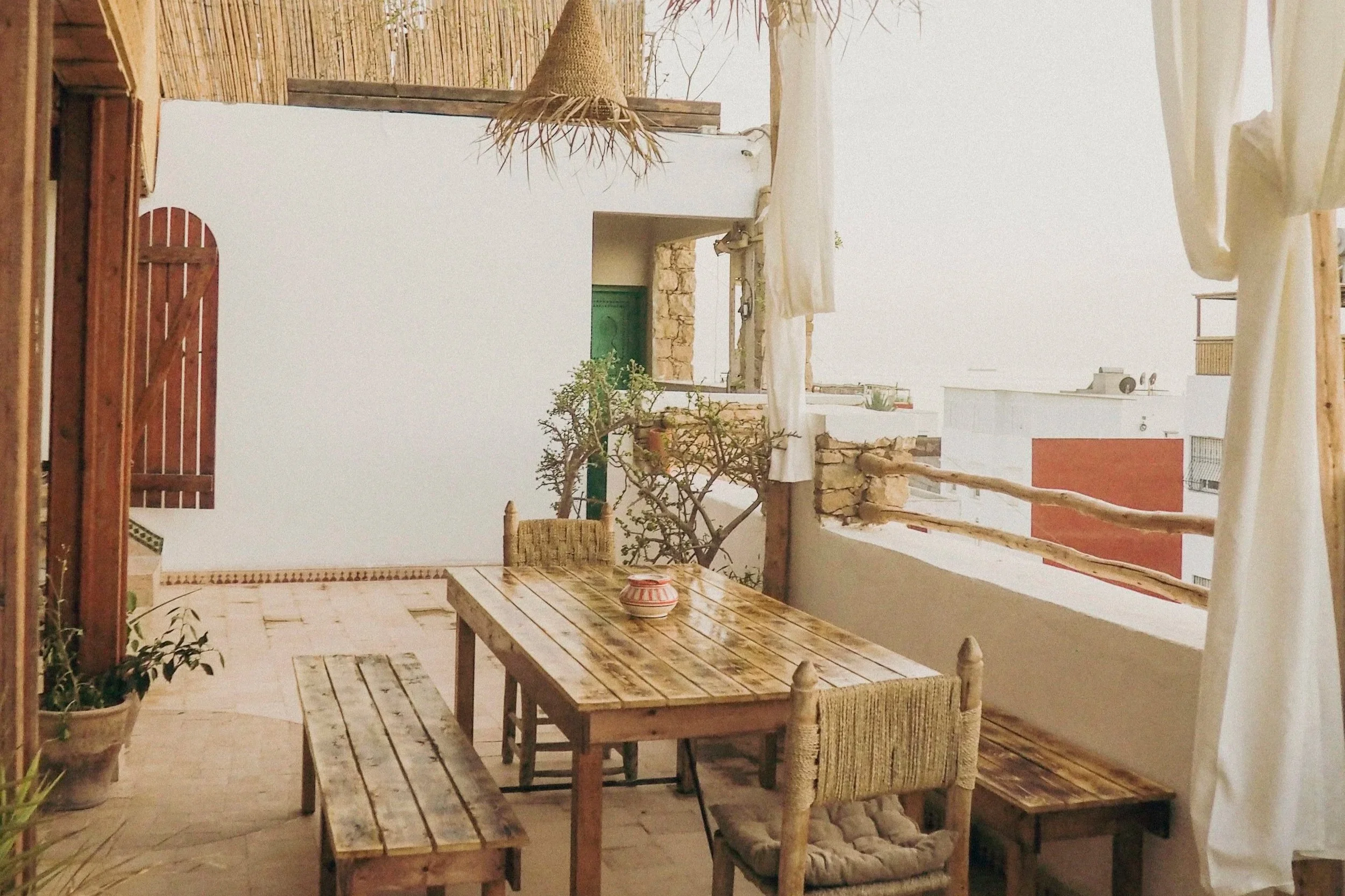 Balcon ensoleillé avec une table en bois, des chaises en bois avec assises en corde, des plantes en pot, un rideau blanc, et une vue partielle sur des bâtiments environnants.